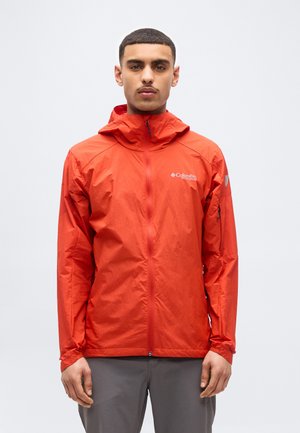 Uomo che indossa una giacca con cappuccio Columbia Titanium di colore arancione vivace, chiusa con cerniera, in piedi davanti a uno sfondo chiaro e uniforme.