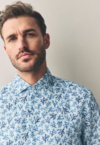 Vaaleansininen napitettava paita, jossa on koralliriuttakuosi tummansinisillä sävyillä. Kangas näyttää sileältä ja paidassa on klassinen kaulus.