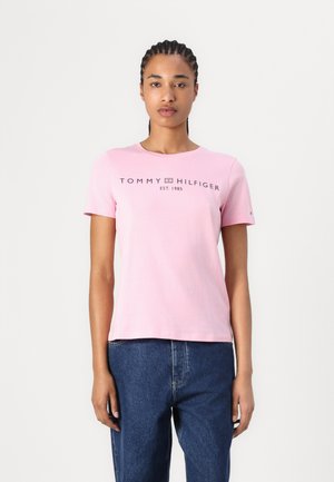 Donna con i capelli intrecciati che indossa una t-shirt rosa chiaro Tommy Hilfiger e jeans blu, in piedi contro uno sfondo bianco semplice.