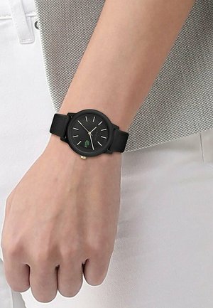 Montre noire avec couronne en or, index blancs et logo crocodile vert portée au poignet gauche sur une peau claire, avec un pantalon blanc et un haut gris.