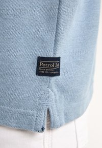 Světle modrá košile s krátkým rukávem vyrobená z texturované látky. Obsahuje námořnickou etiketu s nápisem "Petrol Ltd" a "DENIM DRIVEN" vyšitým zlatou barvou.