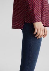 Camicia a fantasia rossa e blu navy con punti tondi, abbinata a jeans skinny blu scuro. Include uno spacco sul bordo della camicia. Mano appoggiata sulla coscia.