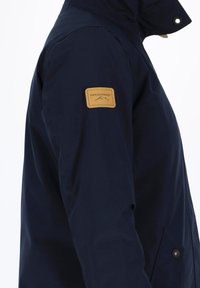 Granatowa kurtka wykonana z gładkiego materiału z brązową skórzaną naszywką na rękawie oraz zapięciem na napy w przedniej kieszeni.