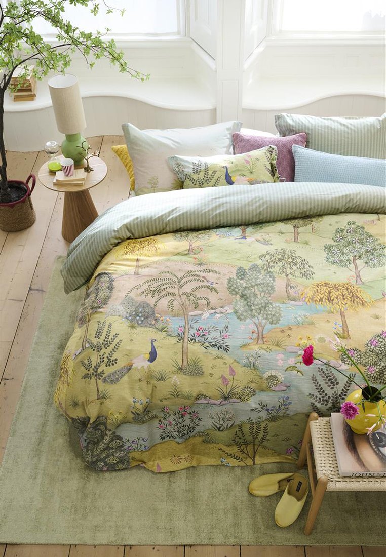 Zestaw pościeli z żółto-zielonym wzorem botanicznym, przedstawiającym drzewa i ptaki. Miękka, teksturowana tkanina z wieloma poduszkami w różnych kolorach i wzorach.