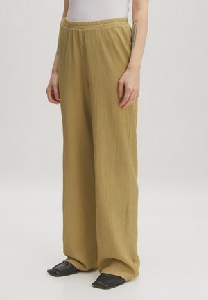 Pantalon large en tissu texturé de couleur vert olive clair, avec une taille élastique, associé à des chaussures noires à bout carré.
