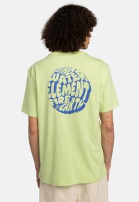 Lys grøn bomulds t-shirt med et stort blåt grafisk design, hvor ordene "VIND", "VAND", "ELEMENT", "ILD" og "JORD" fremstår i et cirkulært mønster.