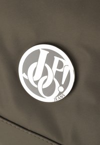 Temno siva tkanina z gladko teksturo vsebuje krožni srebrni logotip s stiliziranim besedilom "JP!" ter besedo "JEANS" na dnu.