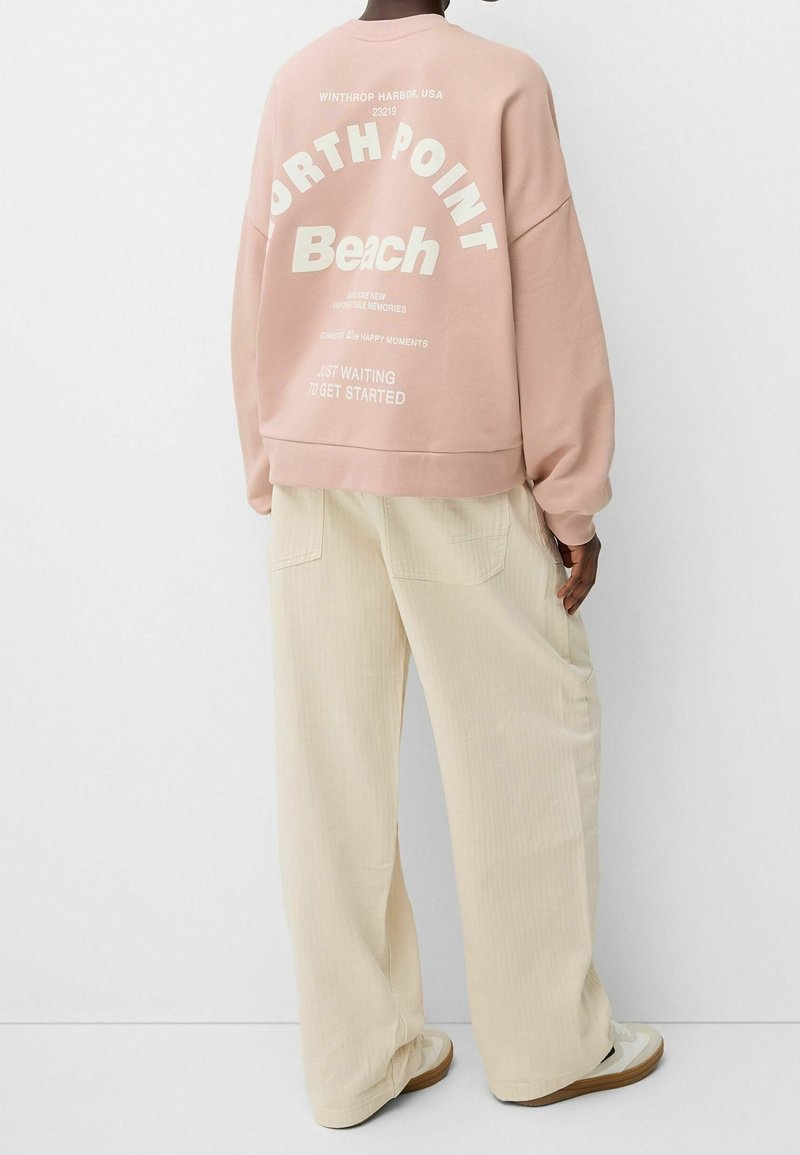 Sweat-shirt rose clair avec texte et graphismes blancs au dos, associé à un pantalon large couleur crème. Tissu doux et coupe décontractée.