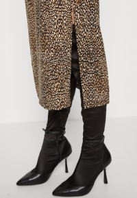 Pitkän leopardikuosiset takki, jossa on edessä halkio, mustien satiinimaisten kireiden housujen ja mustien kärjellä varustettujen stiletto-korkokenkien, joissa on nilkkavetoketjut, kanssa.