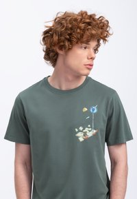 Groene katoenen t-shirt met een grafische print van boeken en een wereldbol op de linkerborst. Ronde halslijn en korte mouwen.