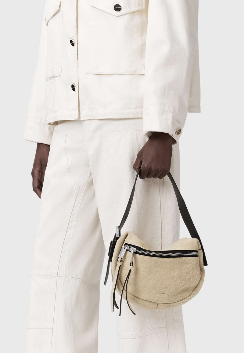 Personne portant une veste et un pantalon écru tenant un petit sac à main en daim beige avec une fermeture éclair et une sangle noires sur le côté.