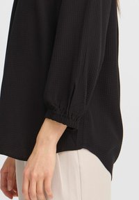 Top nero in tessuto texture con maniche lunghe a sbuffo e polsini elasticizzati, caratterizzato da una vestibilità rilassata e un orlo curvo. Abbinato a pantaloni chiari.