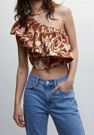 Kvinde iført en brun blomstret enkelt skulder crop-top med flæser og blå højtaljede jeans, med en guldmønt halskæde og hånd i lommen.