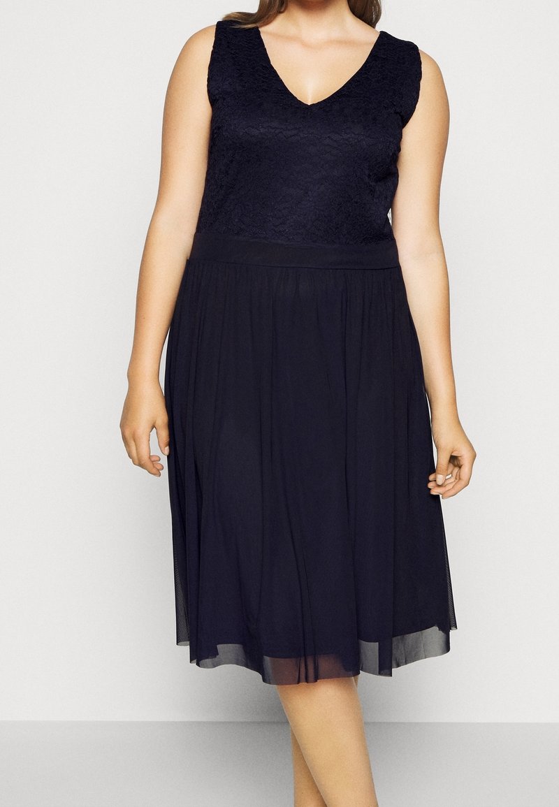 Marineblå klänning med spetsoverdel, V-ringning och en flödande, genomskinlig kjol. Har en åtsittande midja och är ärmlös.