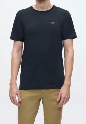 Man wearing a plain black short-sleeve T-shirt with a small chest logo patch and light brown pants, standing against a white background.