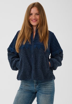Smilende kvinde med langt hår iført en mørkeblå fleecejakke med lynlås, quiltede skuldre og blondekant, kombineret med blå jeans.