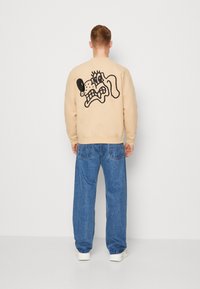 Bežový svetr s velkým černým karikaturálním obličejem na zadní straně. V kombinaci s volnými modrými džíny a bílými teniskami. Hladká textura.