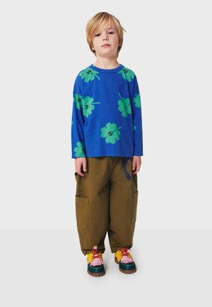 Chemise à manches longues bleue avec un motif floral vert, associée à un pantalon cargo olive avec des poches latérales et des chaussures multicolores avec des accents décoratifs.