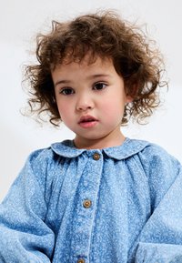 Lyseblå kjole med blomsterprint, rund krave, lange pufærmer og træknapper foran. Blødt, struktureret stof.