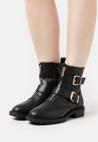 Bottes en cuir noir à la cheville avec deux fermetures éclair latérales, brides ajustables et boucles dorées, dotées d'une semelle en caoutchouc texturée.