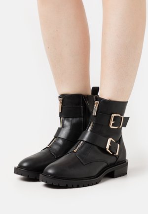 Mustat nahkaiset nilkkurit, joissa on kaksi sivuvetoketjua, säädettävät hihnat ja kullanväriset soljet, teksturoidulla kumipohjalla.