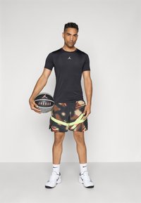 Atlet, der holder en sort og grå Jordan basketball, iført en sort Jordan T-shirt, multifarvede shorts med neonstriber, hvide Nike sneakers og strømper.