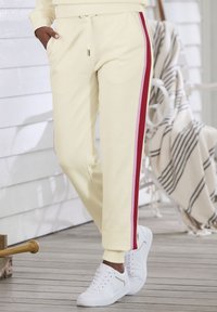 Cremefarbene Jogginghosen mit roten Seitenstreifen, elastischem Bund und Taschen. Kombiniert mit weißen Sneakers mit leichten Akzenten. Holzfußboden im Hintergrund.