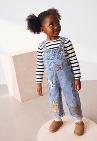 Denim-overalls med frontlomme, dekoreret med farverige dyreapplikationer; båret over en sort-hvid stribet langærmet trøje og brune slip-on sko.