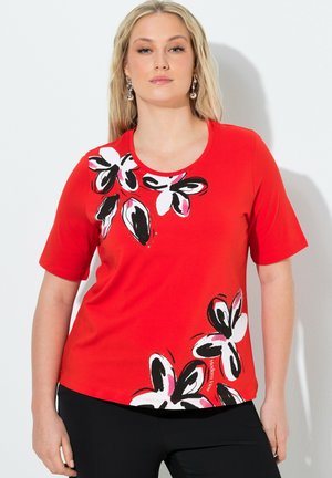 Kobieta w czerwonej koszulce z krótkim rękawem z czarno-biało-różowymi kwiatowymi wzorami na górnej lewej i dolnej prawej stronie.