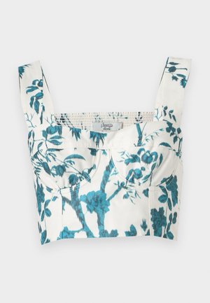 Kvinde iført en hvid og turkisblomstret cropped top med brede stropper, sorte bukser med vide ben og sorte åbne hæle, stående med den ene hånd i lommen.