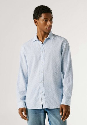 Giovane uomo che indossa una camicia azzurra a fantasia con bottoni e jeans blu, in piedi con la mano destra rilassata, che guarda di lato.