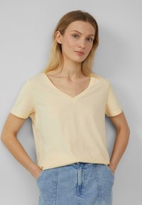 Lichtgele V-hals T-shirt met korte mouwen, gemaakt van zachte stof, gecombineerd met een spijkerrok, weergegeven tegen een neutrale achtergrond.