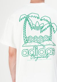 Bela bombažna majica s zelenim grafičnim motivom palm in valov, z napisom "adidas Originals" na dnu.