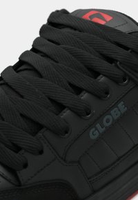 Baskets Black Globe avec des lacets noirs épais, des panneaux en cuir noir et un logo rouge sur la languette.