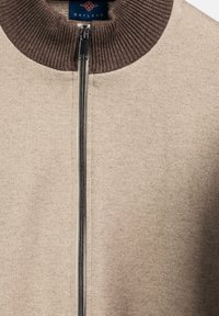 Beige Bomberjacke mit einem braunen Rippkragen und einem durchgehenden Reißverschluss. Der Stoff hat eine strukturierte Oberfläche mit dezenten Sprenkeln.