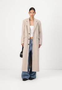 Beige ribbed coat with a long silhouette, notch lapels, and single button closure, paired with wide-leg blue jeans and a black handbag.