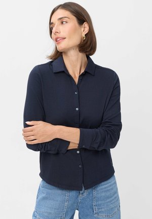 Frau mit kurzen braunen Haaren, die goldene Creolen trägt, ein dunkelblaues Hemd mit Knöpfen und hellblaue Jeans mit aufgesetzten Taschen.