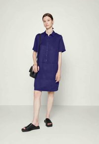 Mulher em pé, vestindo um vestido camisola azul escuro de manga curta com botões, uma bolsa preta cruzada e sandálias de plataforma abertas de dedo preto, em um fundo liso.