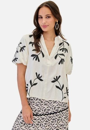 Mujer con blusa blanca bordada con hojas negras y falda a juego con estampado de hojas negras, complementada con grandes pendientes de aro.