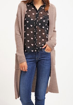 Kvinde iført en lang beige strikket cardigan over en sort, gennemsigtig bluse med pink hjerter og blå jeans, stående med den ene hånd i lommen.