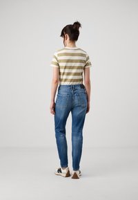 Randig olivgrön och krämfärgad t-shirt instoppad i blå denimjeans med smal passform. Har bakfickor och stygn detaljer. Neutral bakgrund.