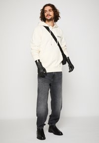 Krēma kapuci džemperis ar priekšējo kabatu, kombinēts ar pelēkiem džinsiem. Aksesuāri ietver melnu crossbody somu un melnas potītes zābakus.