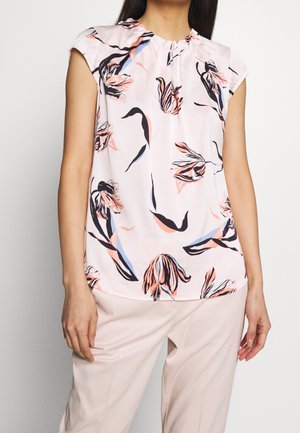 Kobieta ubrana w różową, bezrękawną bluzkę w czarno-brzoskwiniowo-niebieski kwiatowy wzór oraz różowe spodnie.