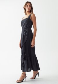 Black maxi dress with a square neckline, ruffled hem, and tie waist detail. Features a flowing silhouette and soft texture. Pair of black high-heeled sandals.