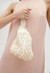 Biała torebka o teksturowanej powierzchni z frędzlami ozdobionymi koralikami, z plecionym uchwytem. Torebka jest trzymana przez osobę ubraną w jasnoróżową sukienkę.