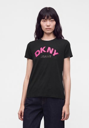 Jeune femme aux longs cheveux foncés portant un t-shirt noir DKNY Jeans et un pantalon sombre, debout devant un fond blanc uni.