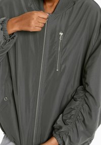 Šedá bomber bunda vyrobená z hladkého materiálu, s žebrovaným límcem, zipem vpředu, postranními kapsami a nařasenými detaily na rukávech.