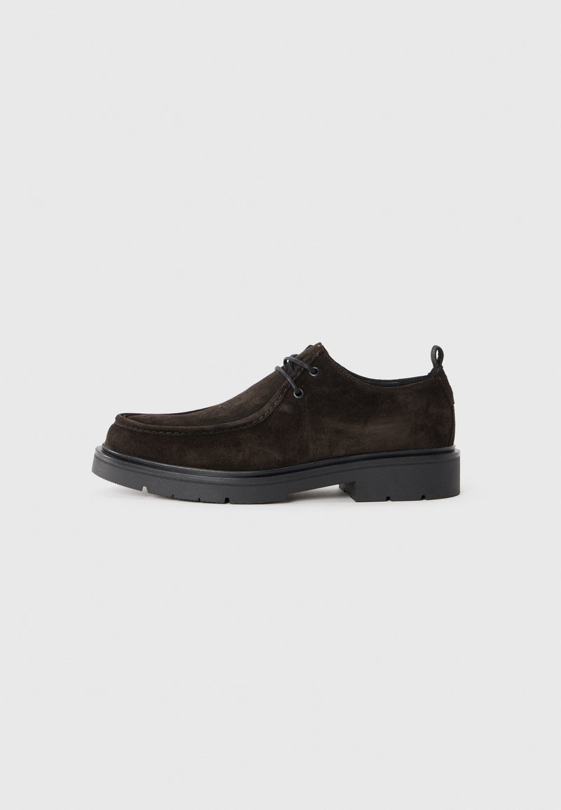 Zapato de ante marrón oscuro con cordones, suela gruesa negra y tirador, sobre fondo gris, vista lateral.