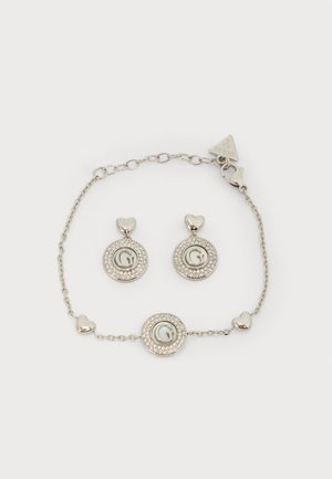 Bracciale tono argento e orecchini abbinati con dettagli a forma di cuore e loghi circolari incastonati con piccoli cristalli.