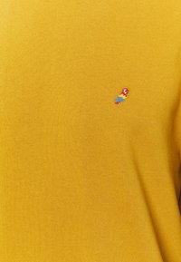 Tessuto giallo senape con un piccolo ricamo colorato a forma di pappagallo nella zona superiore del petto.